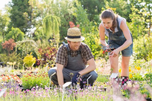Two individuals working together in a well-maintained garden during daylight hours, with lush greenery including trees and shrubs in the background. The foreground features a diverse array of colorful flowers, bordered by a neat grass lawn with a rich green hue, and a paved or gravel pathway partially visible on the right side. The person on the left, squatting, is wearing a straw hat and a checked shirt, and appears to be tending to the flower bed, while the person on the right, standing, is dressed in a sleeveless top and denim shorts, holding garden shears, possibly trimming or pruning plants. The garden appears to be part of an outdoor residential or community space, showcasing a blend of natural plant textures and vibrant blooms, suggesting professional gardening or landscaping services in Earl's Court. The weather looks bright with natural sunlight illuminating the scene, indicating a clear day suitable for outdoor gardening activities. The environment is tidy, organized, and thriving, reflecting ongoing maintenance and care, consistent with gardening and landscaping service standards in the local area.