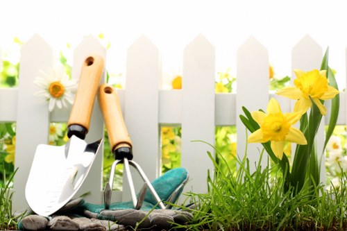 The image showcases an outdoor garden scene in a residential setting, featuring a white picket fence in the background, with a variety of plants and flowers in the foreground. On the ground, there is a patch of lush, green grass interspersed with soil and small plants. Bright yellow daffodils with green leaves are prominently positioned to the right, adding vibrant colour against the natural tones of the garden. To the left, a few white daisies with yellow centers are visible, contributing to the cheerful garden atmosphere. Leaning against the fence are two gardening tools: a small hand trowel with a white blade and a wooden handle, and a cultivator with a green handle, both suggesting ongoing gardening or maintenance activities. The lighting appears natural and bright, indicating a sunny day, which highlights the vivid colours and textures of the plants and tools, reinforcing the outdoor gardening environment. This scene represents a well-tended garden space suitable for professional gardening and landscaping services, such as hedge trimming or lawn care, provided by companies like Hedge Trimming Earls Court, serving gardens in the local area near Earl's Court, SW5 postcode.